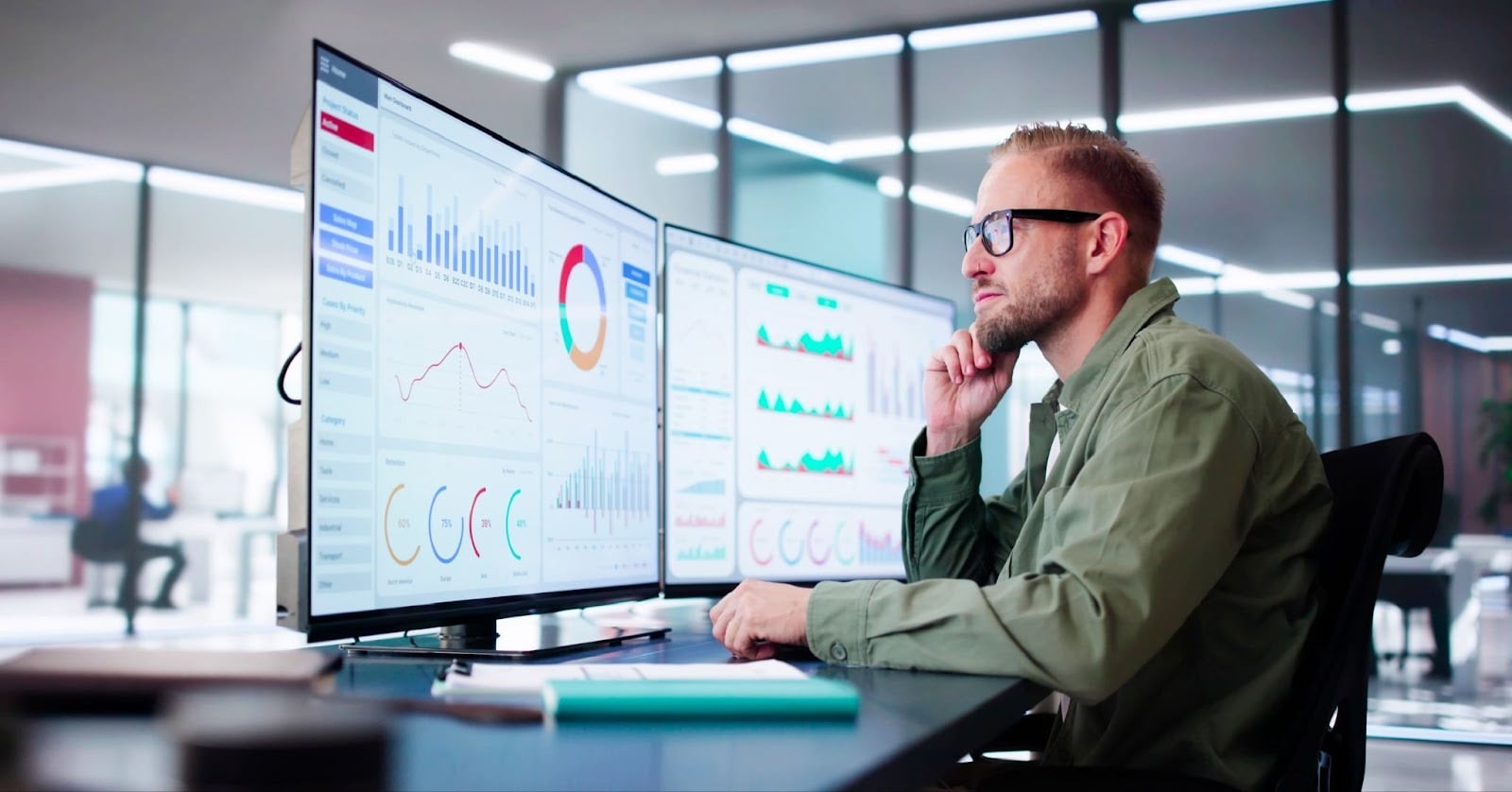 Man with glasses sitting at a desk reviewing colorful analytics dashboards on dual monitors in a modern office
