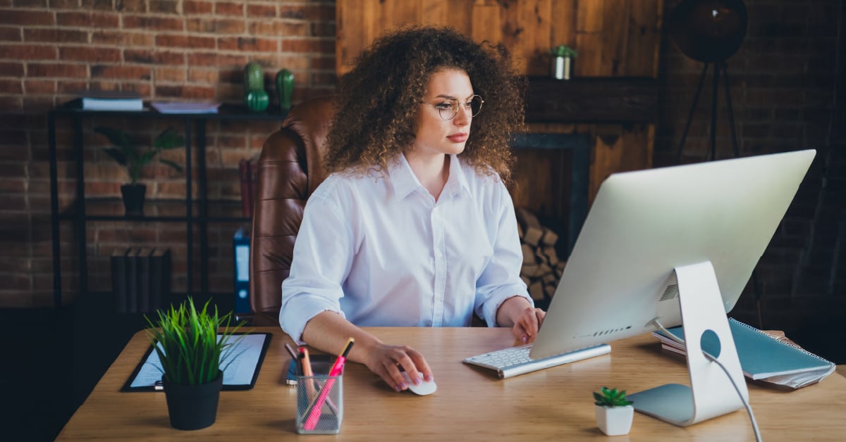 Woman with curly hair working at a desk with a desktop computer in a rustic brick-walled office