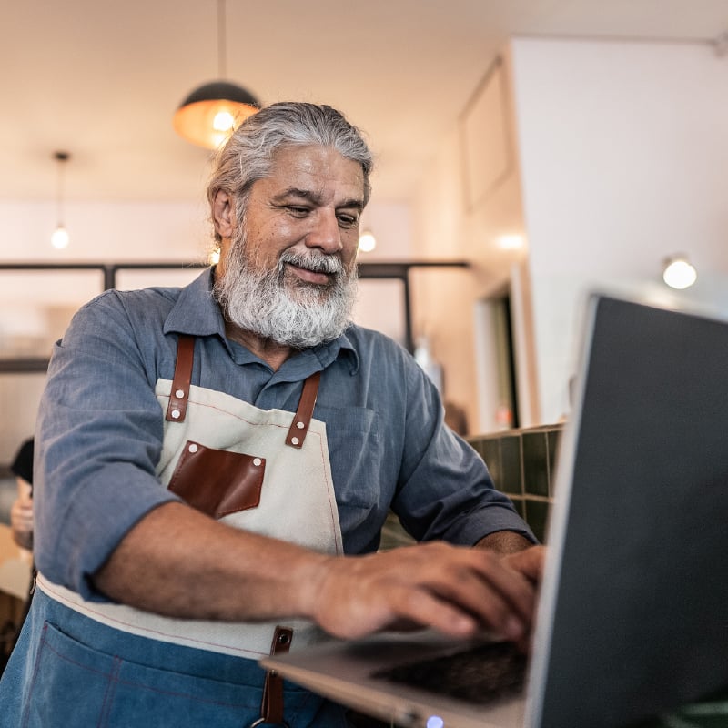 Smiling bearded man wearing a denim shirt and canvas work apron, standing in a workshop or shop setting